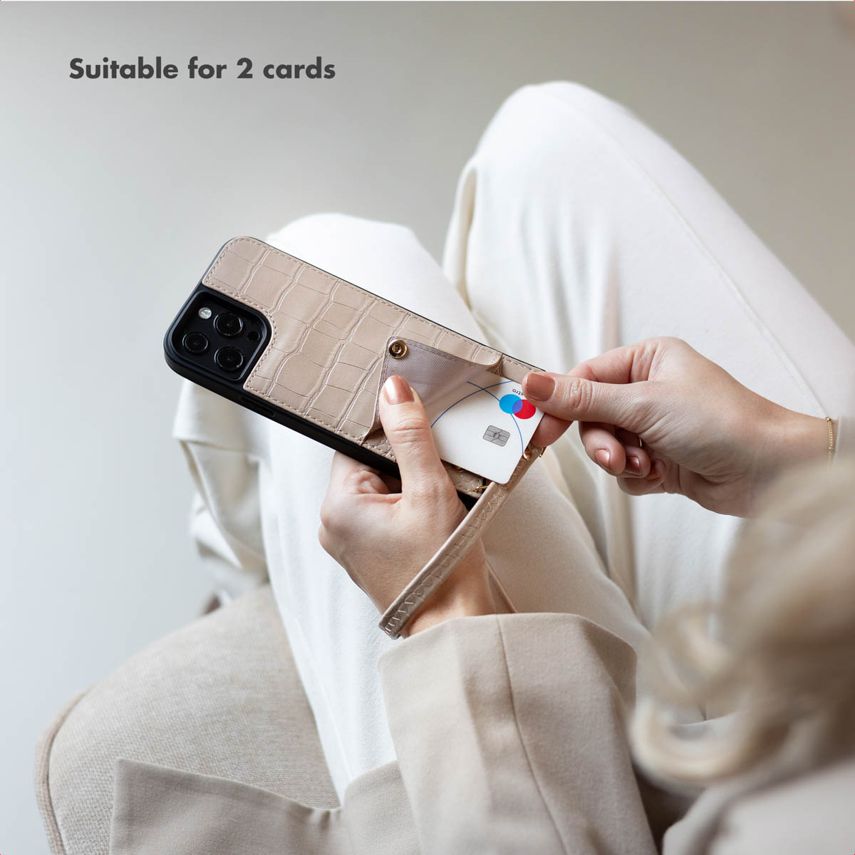 Una mujer con ropa blanca sostiene una funda de teléfono beige con tarjetero y correa.
