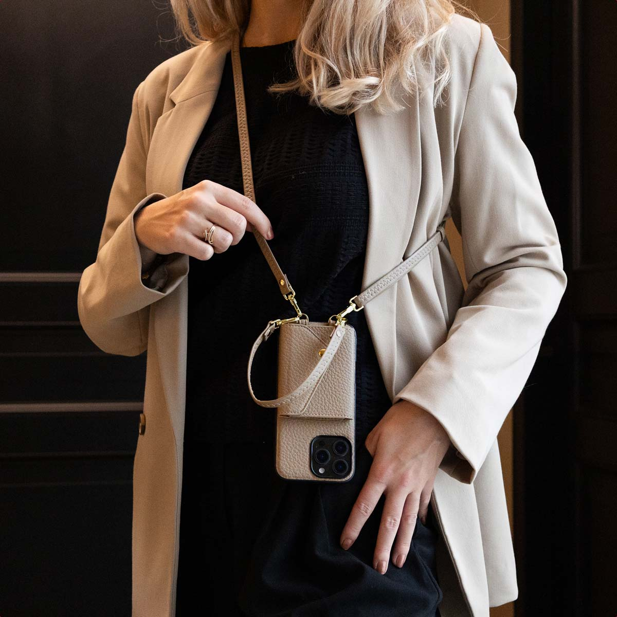 Una donna con top nero e giacca beige indossa una custodia per telefono beige con tracolla.
