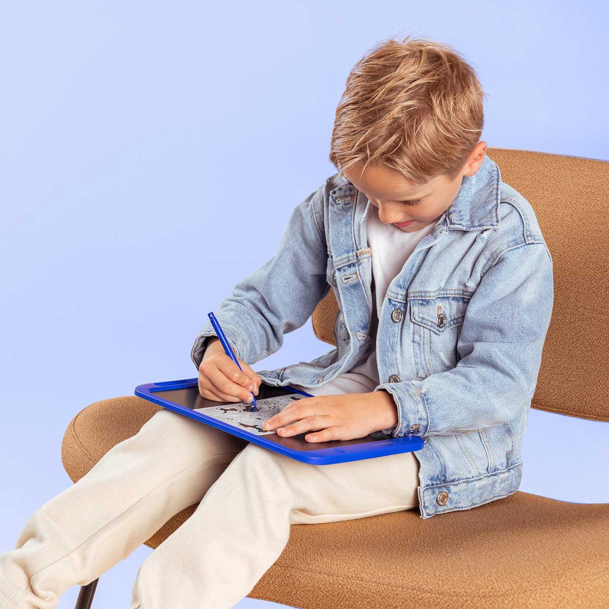 Un bambino biondo in giacca di jeans disegna su un tablet blu seduto su una sedia marrone.