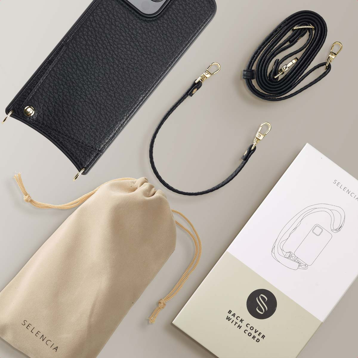 Funda negra para teléfono, dos correas negras, bolsa beige y caja de producto blanca sobre superficie beige.