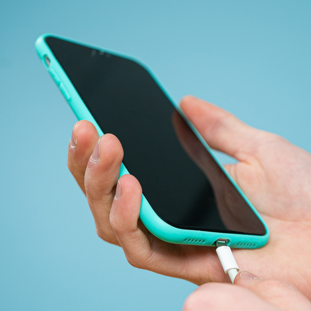 Une main tient un smartphone turquoise tout en branchant un câble de charge blanc sur fond bleu clair.