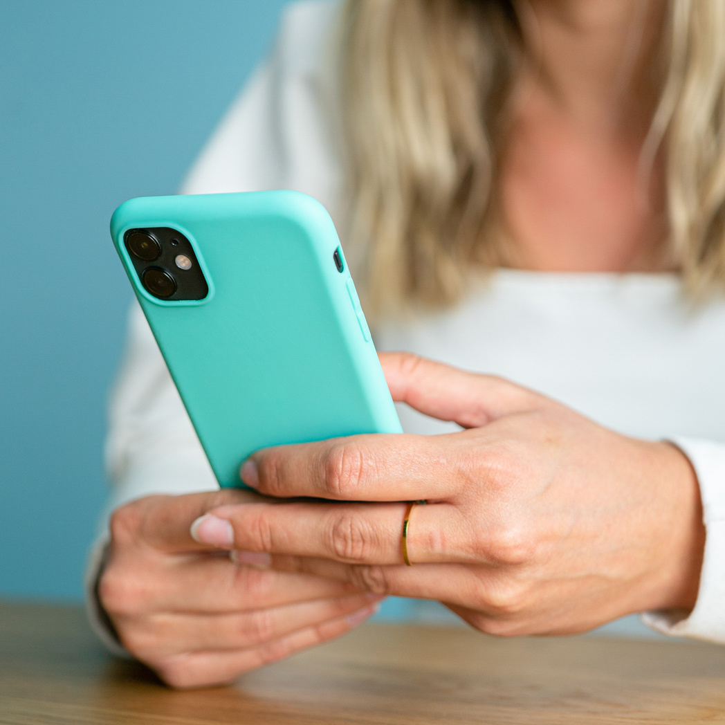 Une femme tient un smartphone avec coque turquoise, assise à une table en bois devant un mur bleu.