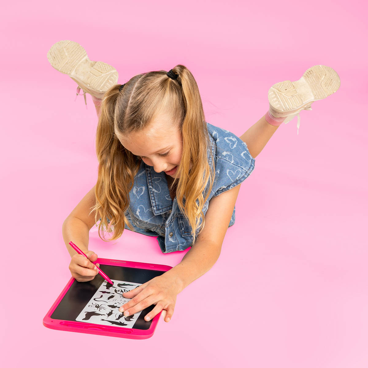 Una bambina bionda sdraiata su sfondo rosa disegna con una penna rosa su uno stencil.