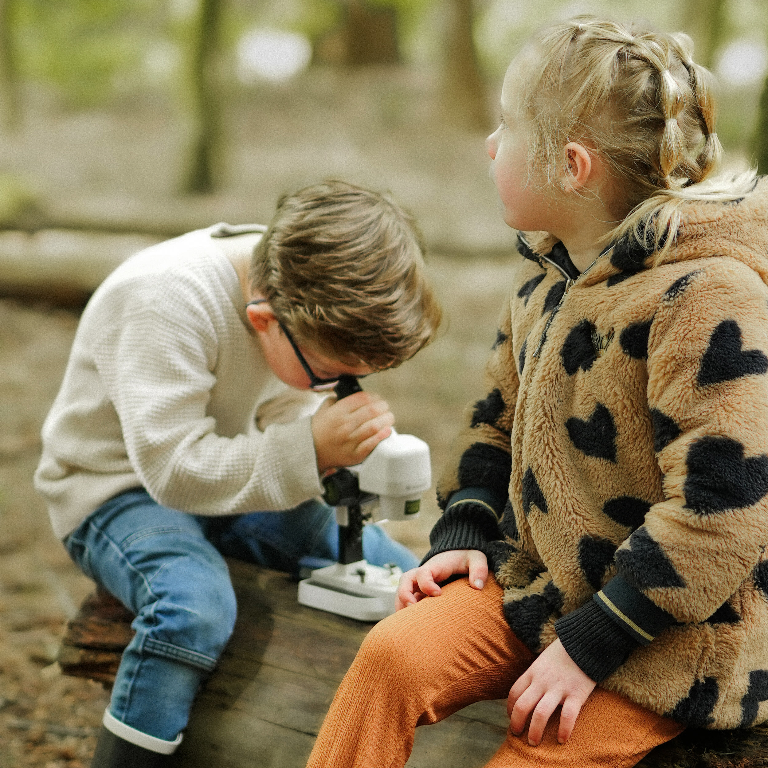 Un niño mira a través de un microscopio mientras una niña se sienta a su lado, al aire libre en un bosque.