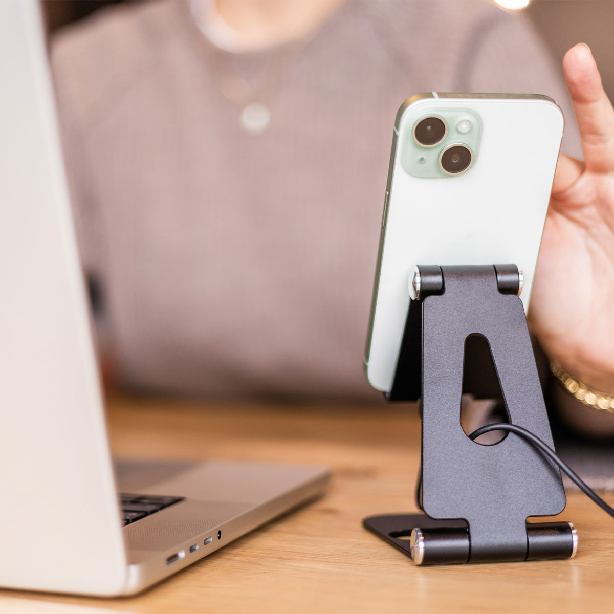Uno smartphone su un supporto nero accanto a un laptop su una scrivania di legno, con una persona sullo sfondo.