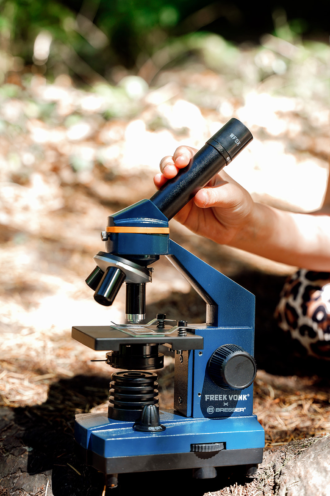 Una mano ajusta el ocular de un microscopio Bresser azul sobre un suelo de bosque.