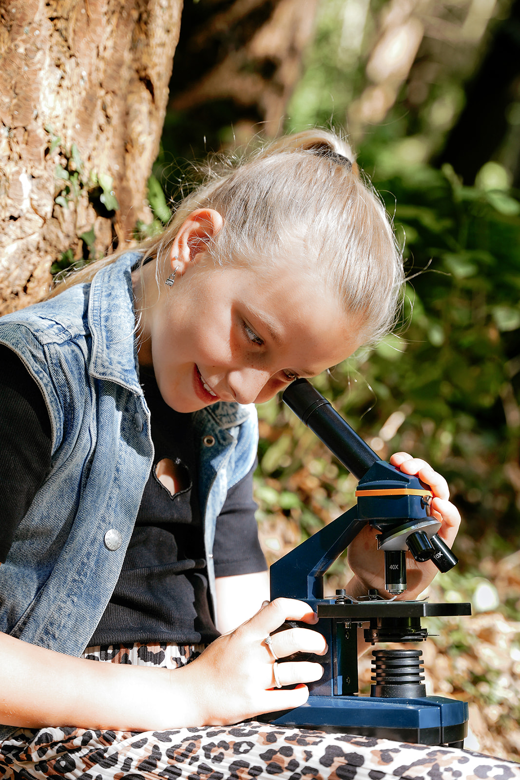 Una niña rubia con chaleco de mezclilla usa un microscopio al aire libre junto a un árbol.