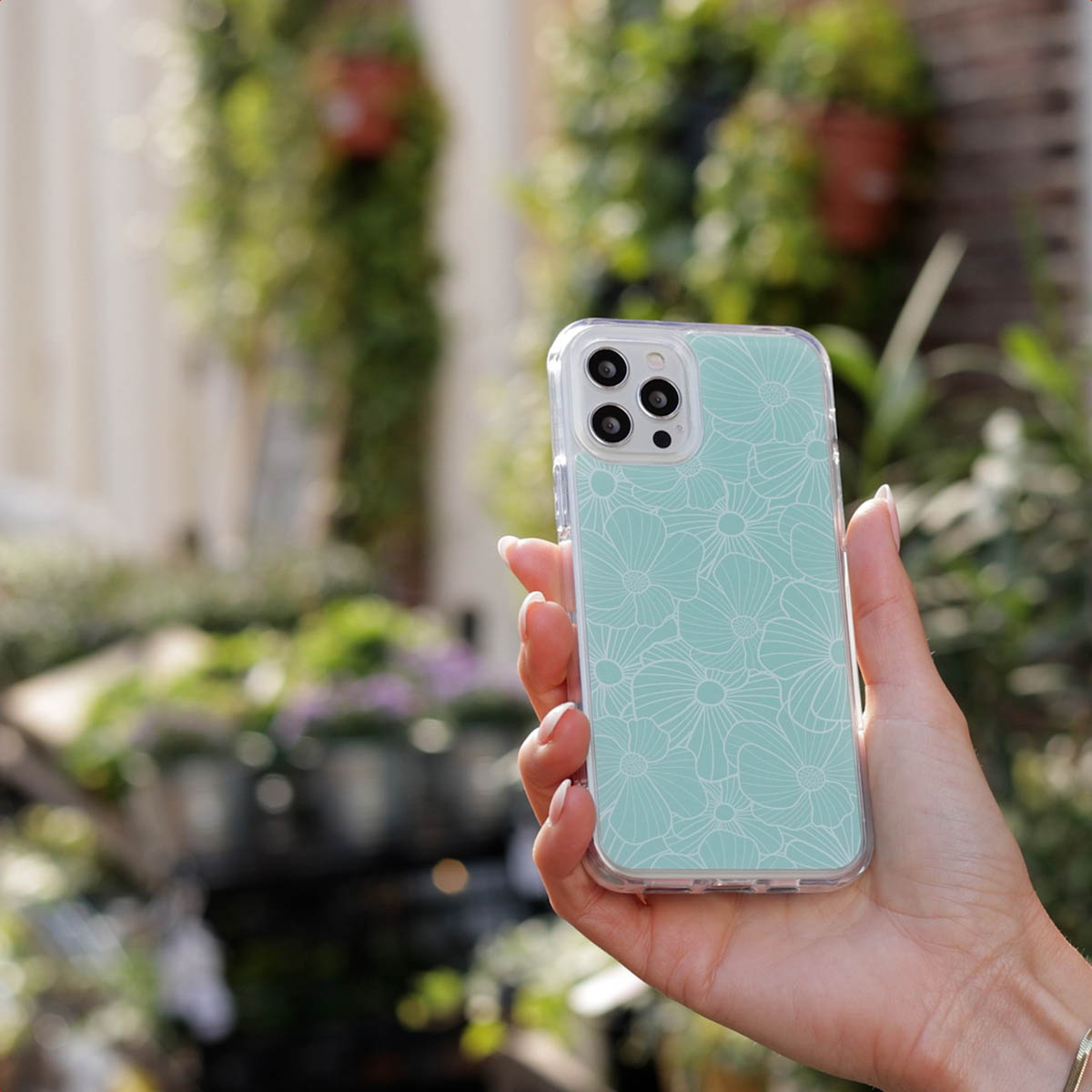 Una mano sostiene un teléfono con funda verde menta y diseño floral blanco frente a un jardín borroso.