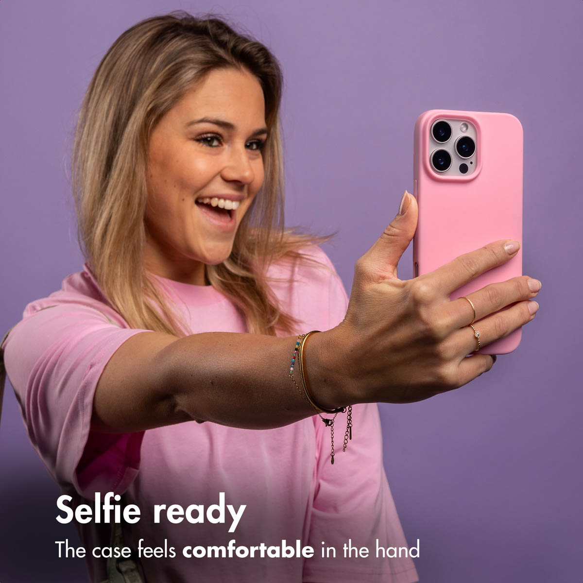 Blond kobieta w różowej koszulce uśmiecha się, robiąc sobie selfie różowym telefonem na fioletowym tle.