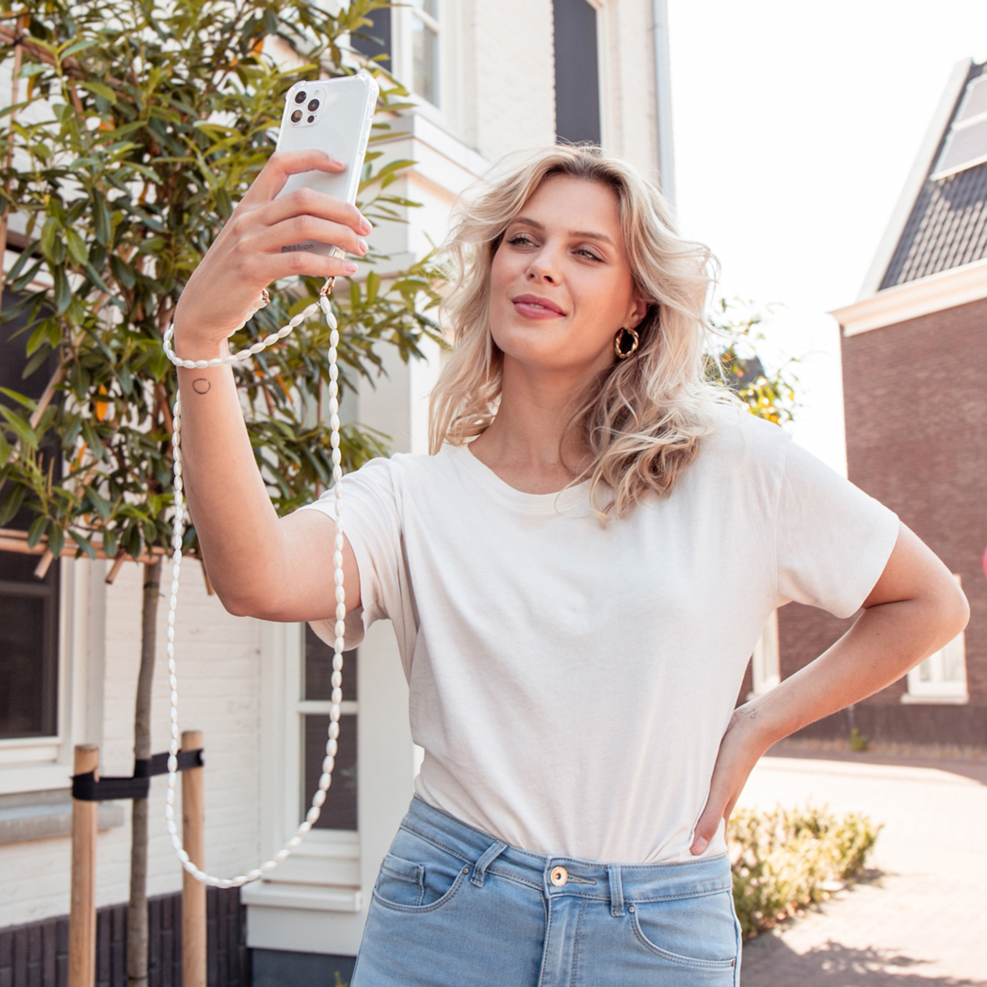 Una mujer rubia con camiseta blanca y jeans se toma un selfie con su teléfono blanco al aire libre frente a un edificio.