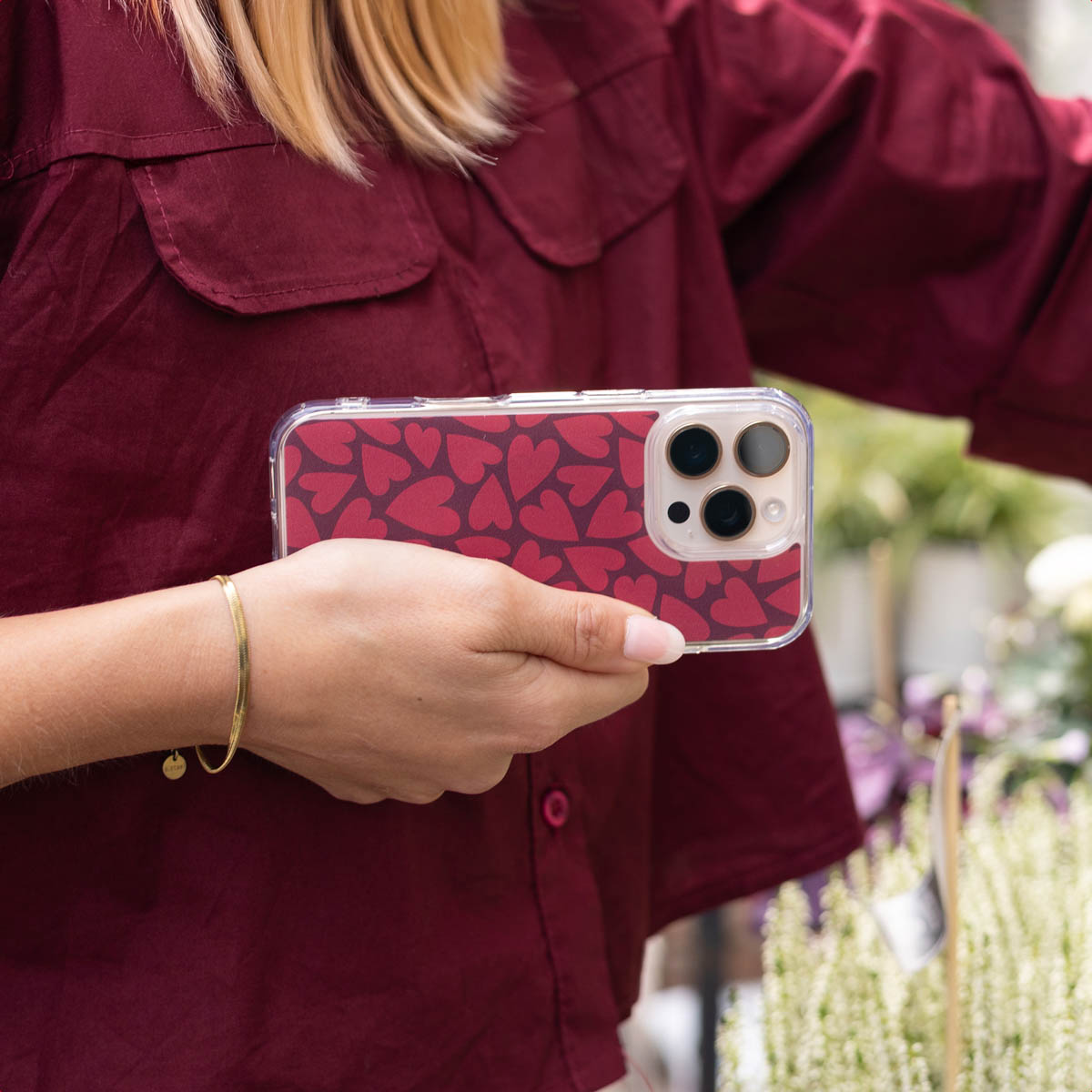 Una persona con camisa granate sostiene un smartphone con funda de corazones rojos al aire libre.