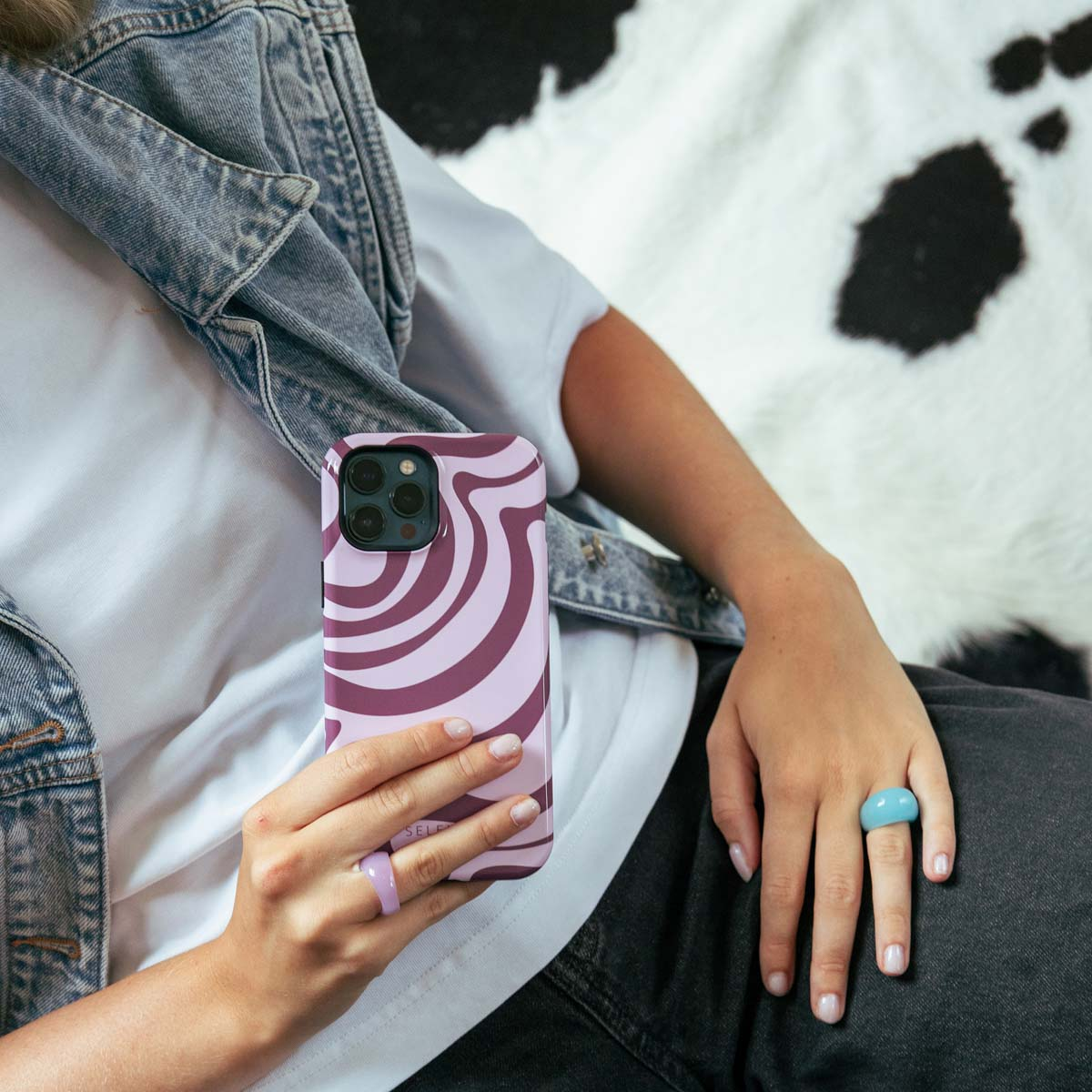 Persona con cellulare viola ondulato, gilet di jeans, camicia bianca e anelli colorati.