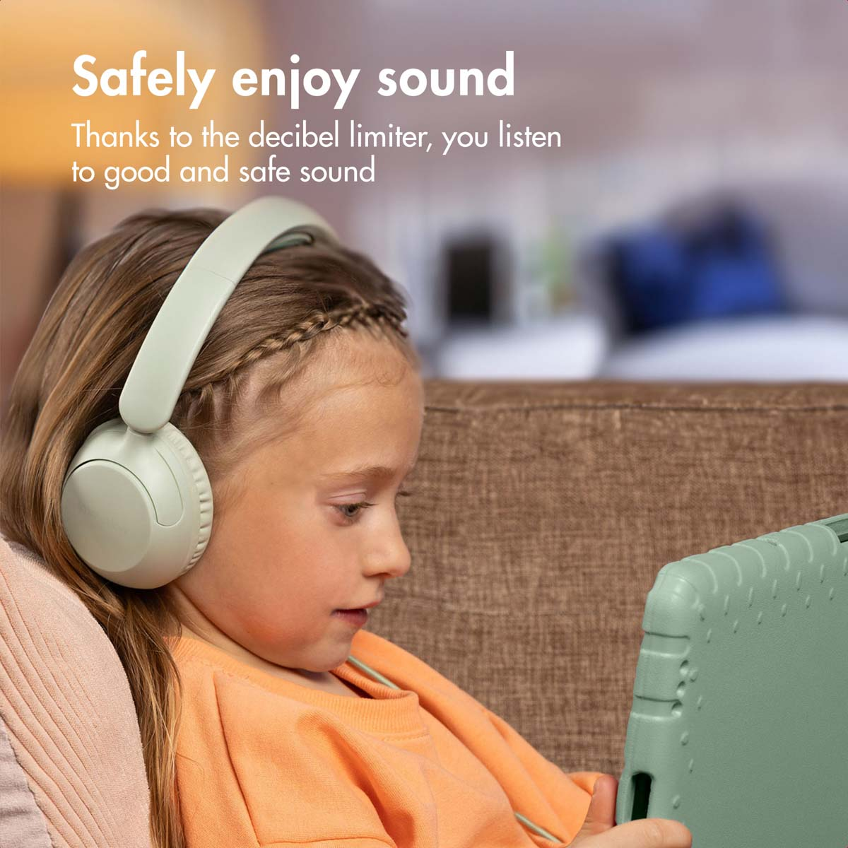 Una niña con camisa naranja lleva auriculares verdes y mira una tableta verde en un sofá.