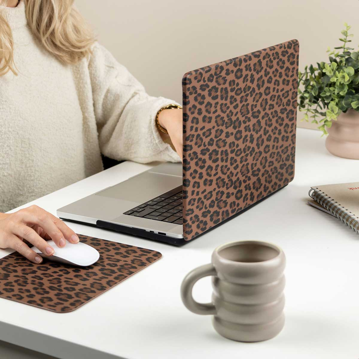 Una mujer usa una laptop con estampado de leopardo y ratón en un escritorio blanco con una taza y una planta.