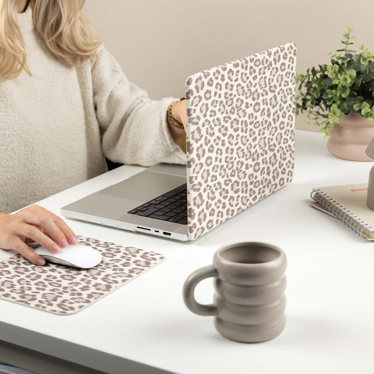 Una mujer escribe en un portátil en un escritorio blanco con accesorios de leopardo y una taza de café.