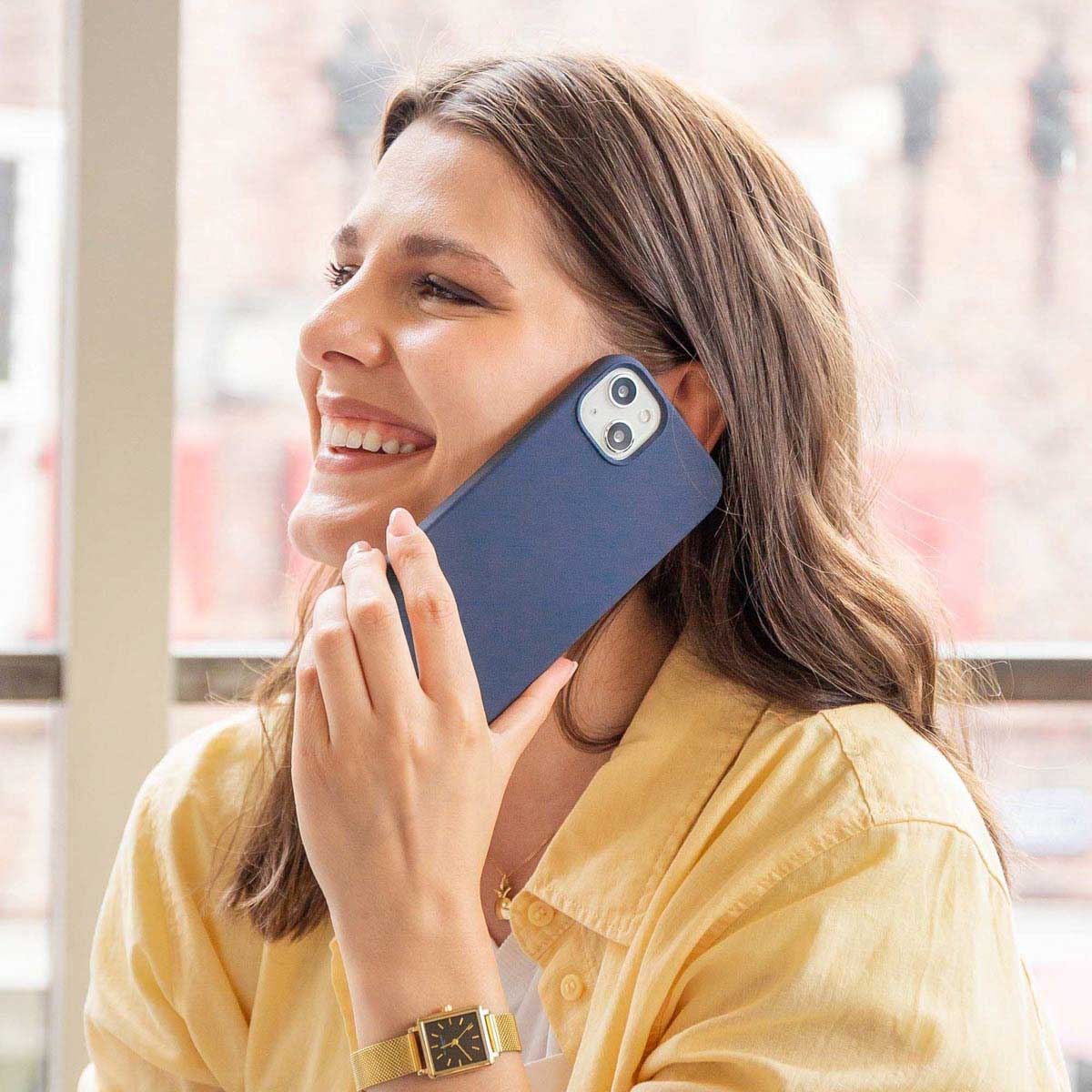 Uśmiechnięta kobieta w żółtej koszuli przykłada do ucha niebieski smartfon.