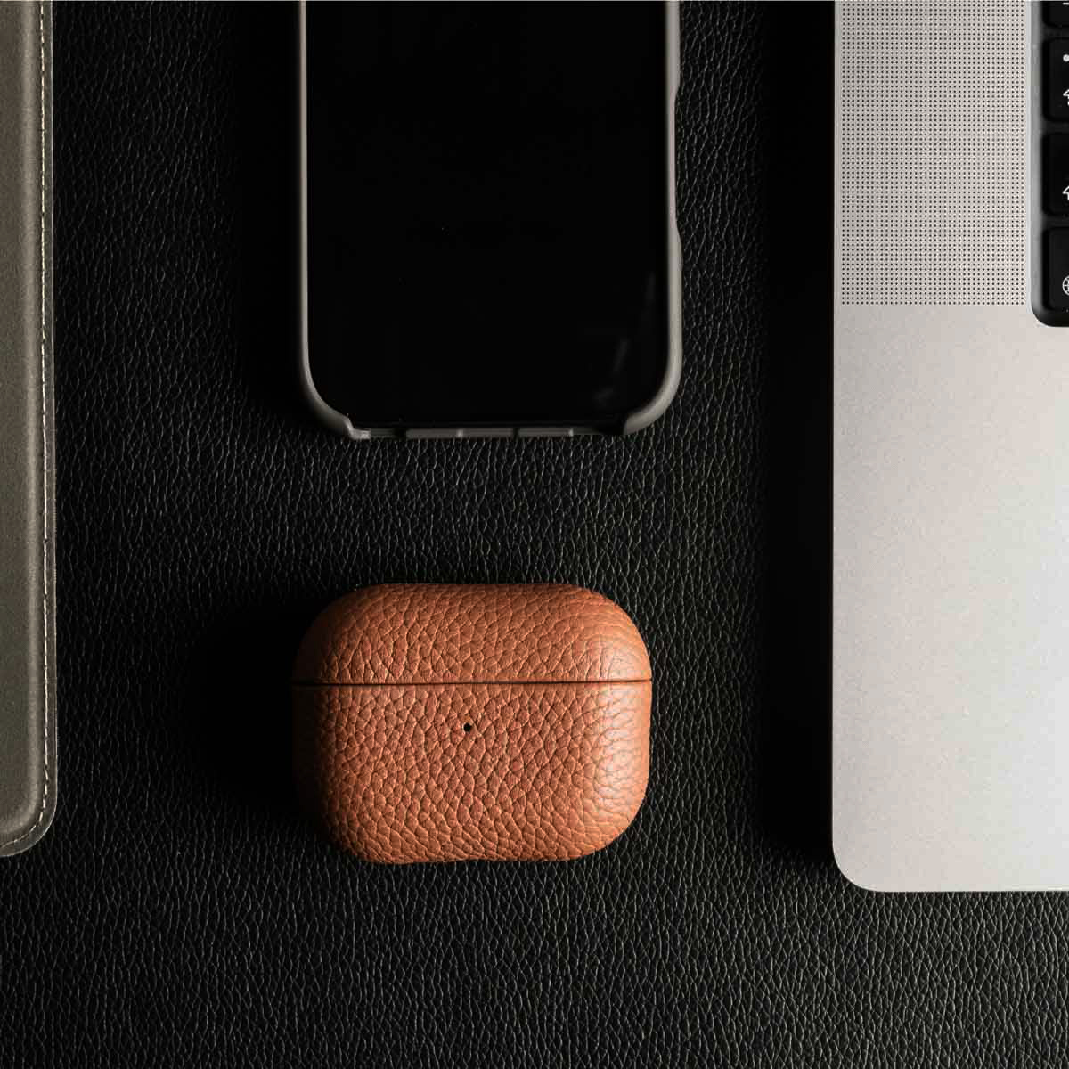 Una custodia per AirPods in pelle marrone si trova accanto a uno smartphone e un laptop su una superficie in pelle nera.