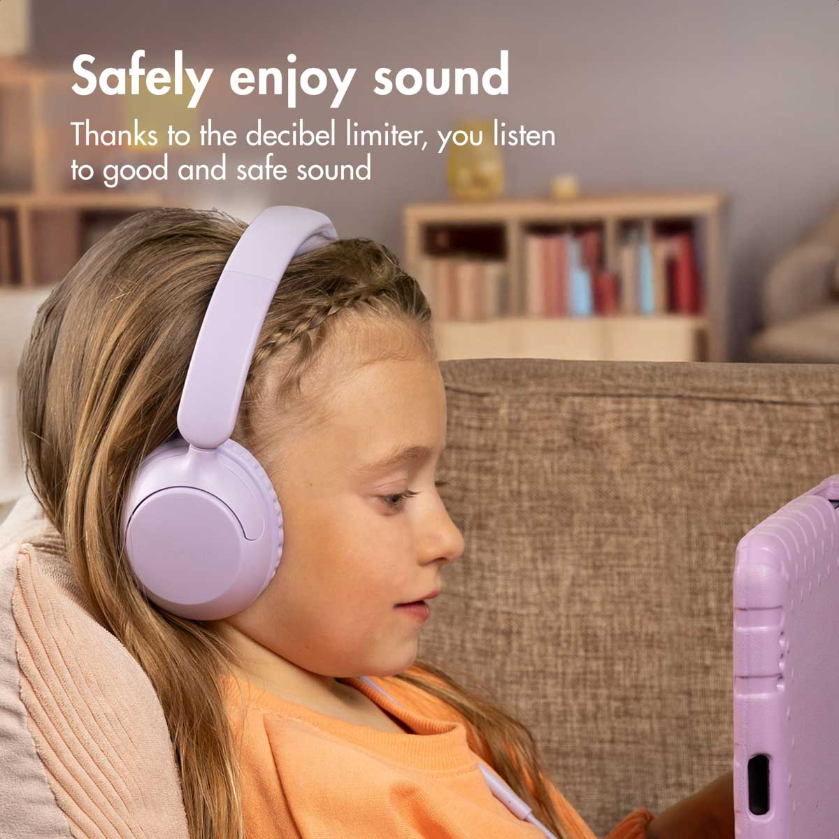 Una niña pequeña con auriculares morados mira una tableta en una habitación acogedora.