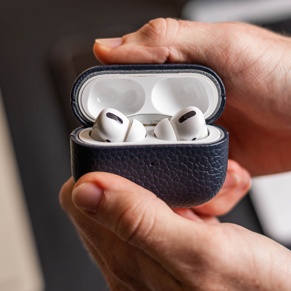 Le mani tengono una custodia in pelle blu scuro aperta contenente due auricolari wireless bianchi.