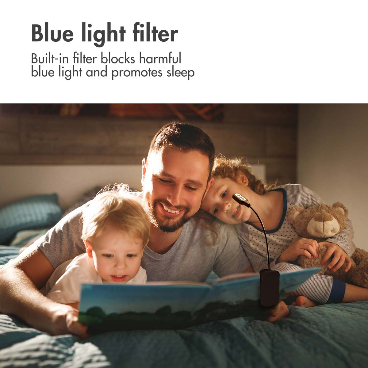Ein Vater und zwei Kinder lesen im Bett ein Buch, beleuchtet von einer kleinen Klemmleuchte.