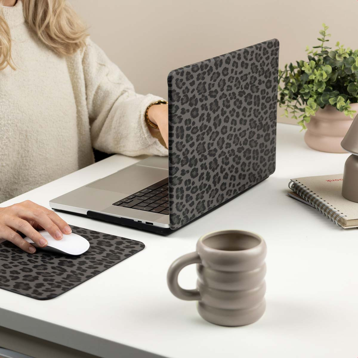 Una persona con suéter blanco usa una laptop y un ratón con estampado de leopardo en un escritorio con taza y planta.
