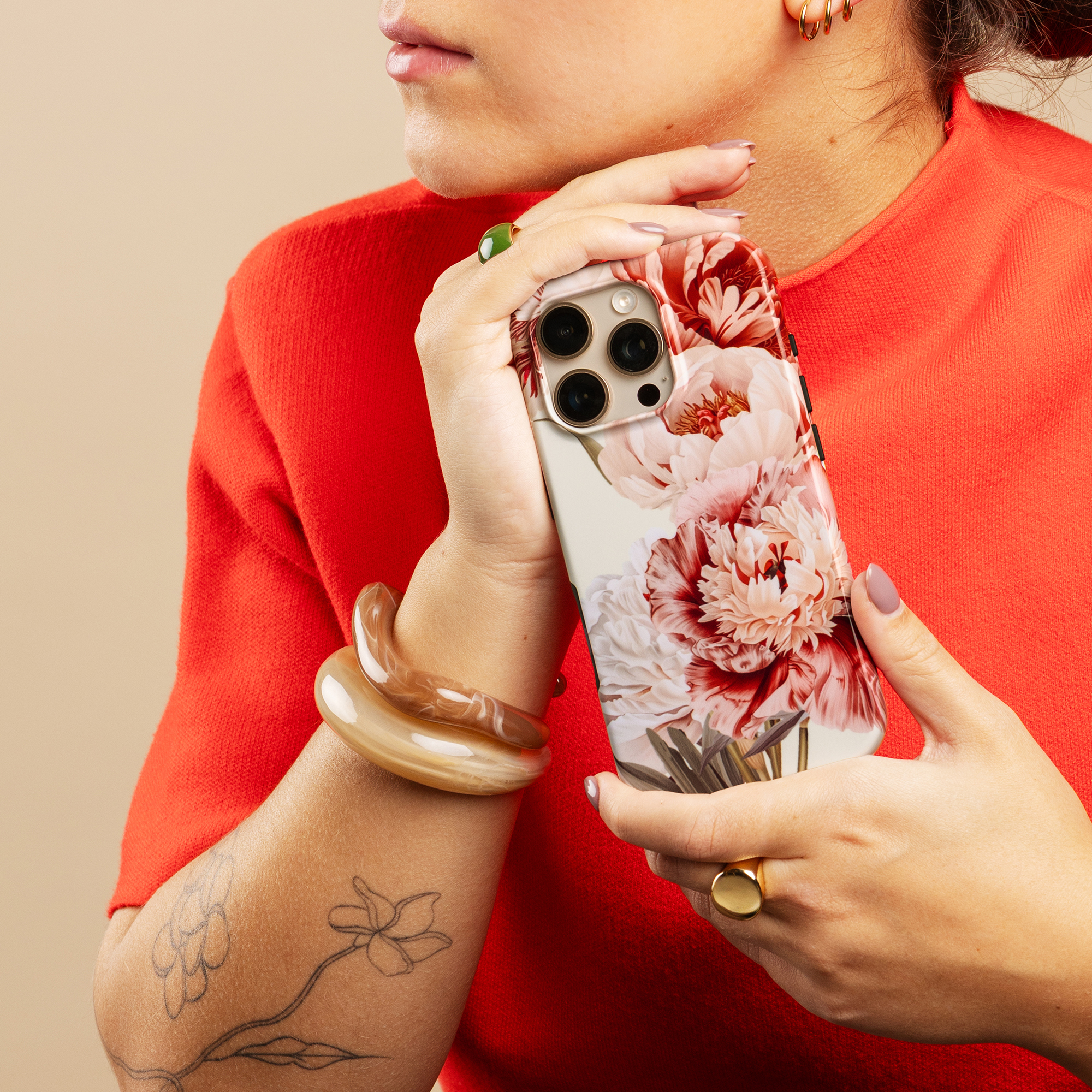Una persona con camisa roja sostiene un teléfono con funda floral, luciendo pulseras y anillos.