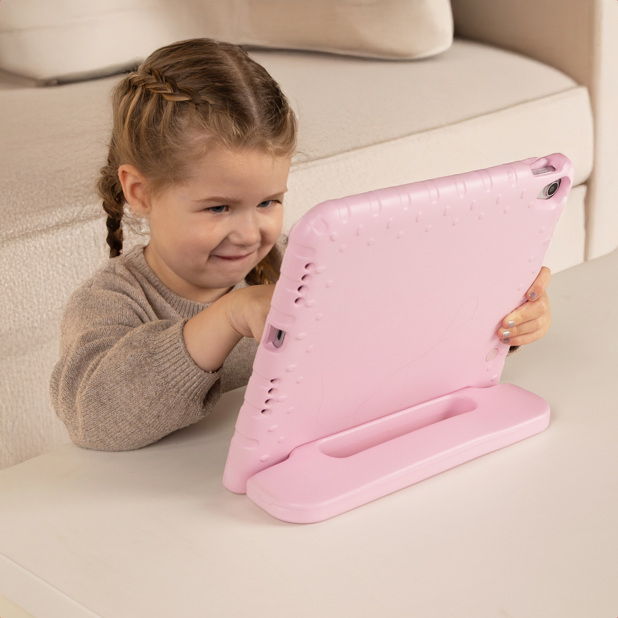 Een jong meisje met vlechten zit aan tafel en gebruikt een tablet in een roze hoes.
