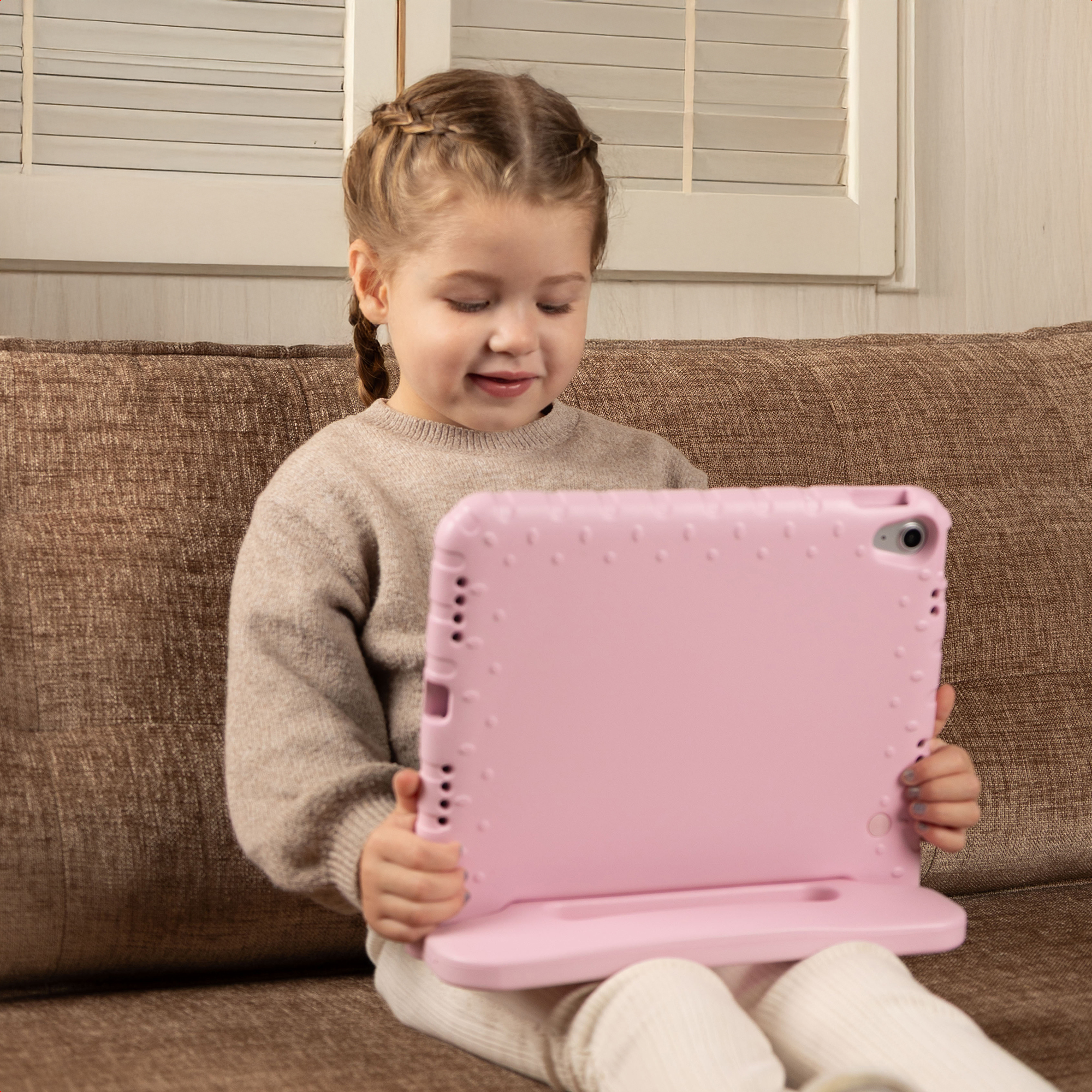 Een jong meisje met gevlochten haar zit op een bank en houdt een grote roze tablet vast.