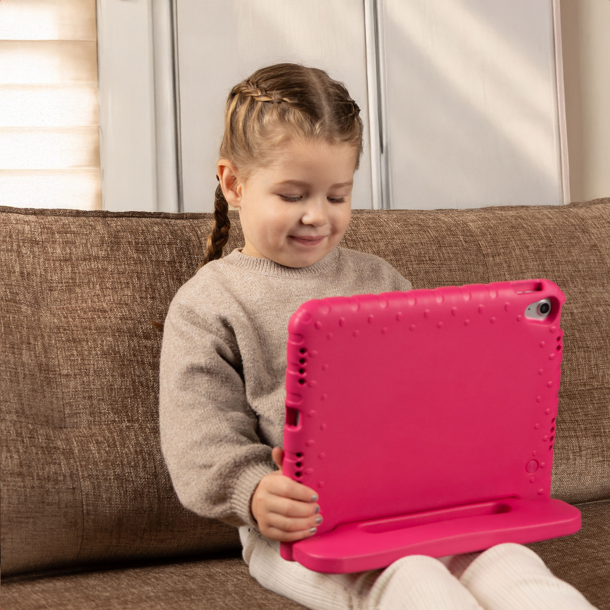 Una bambina con le trecce siede su un divano marrone, usando un tablet in una custodia rosa.