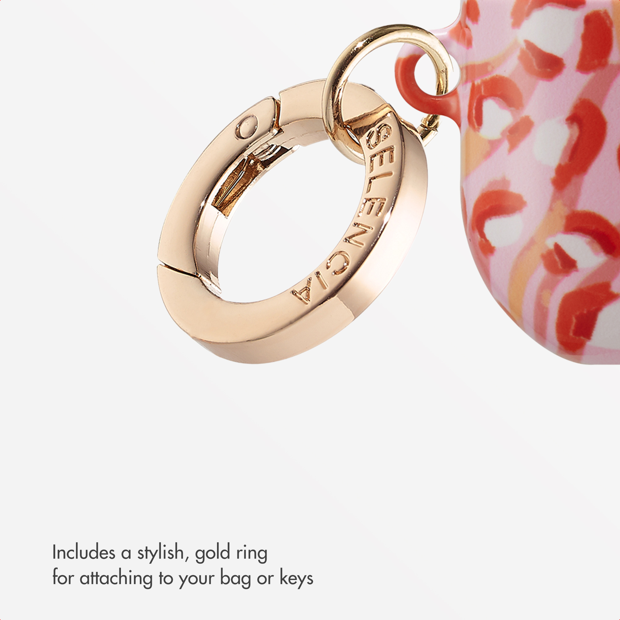 Un anillo dorado con grabado cuelga de un estuche estampado en rosa y blanco sobre fondo blanco.