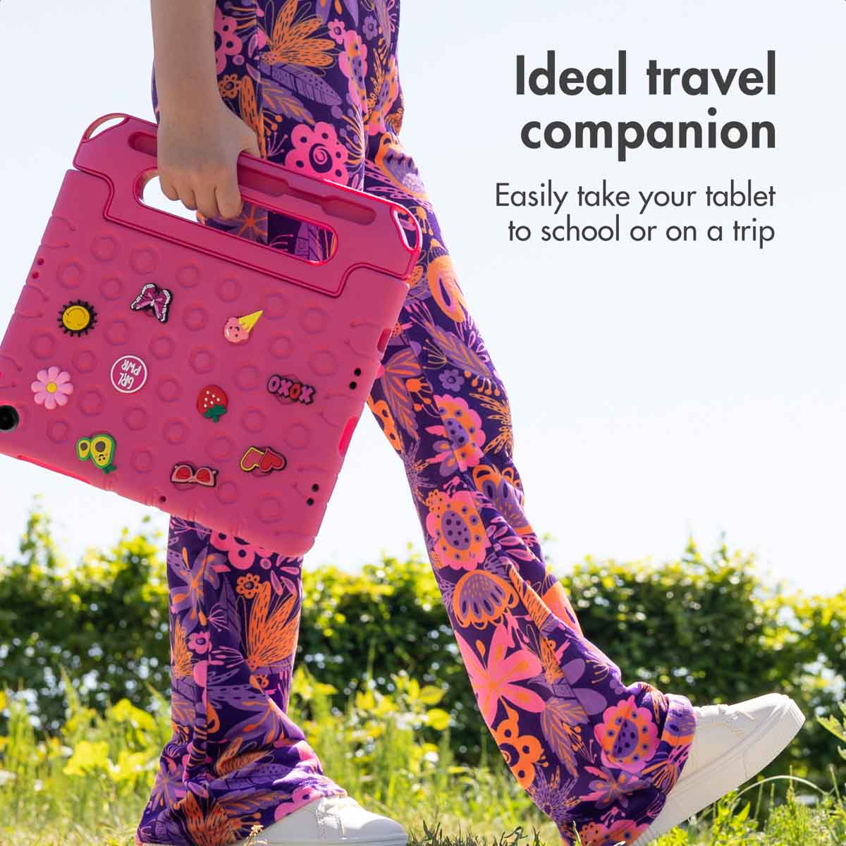 A person walks outdoors in floral pants, carrying a pink protective tablet case with cute stickers.