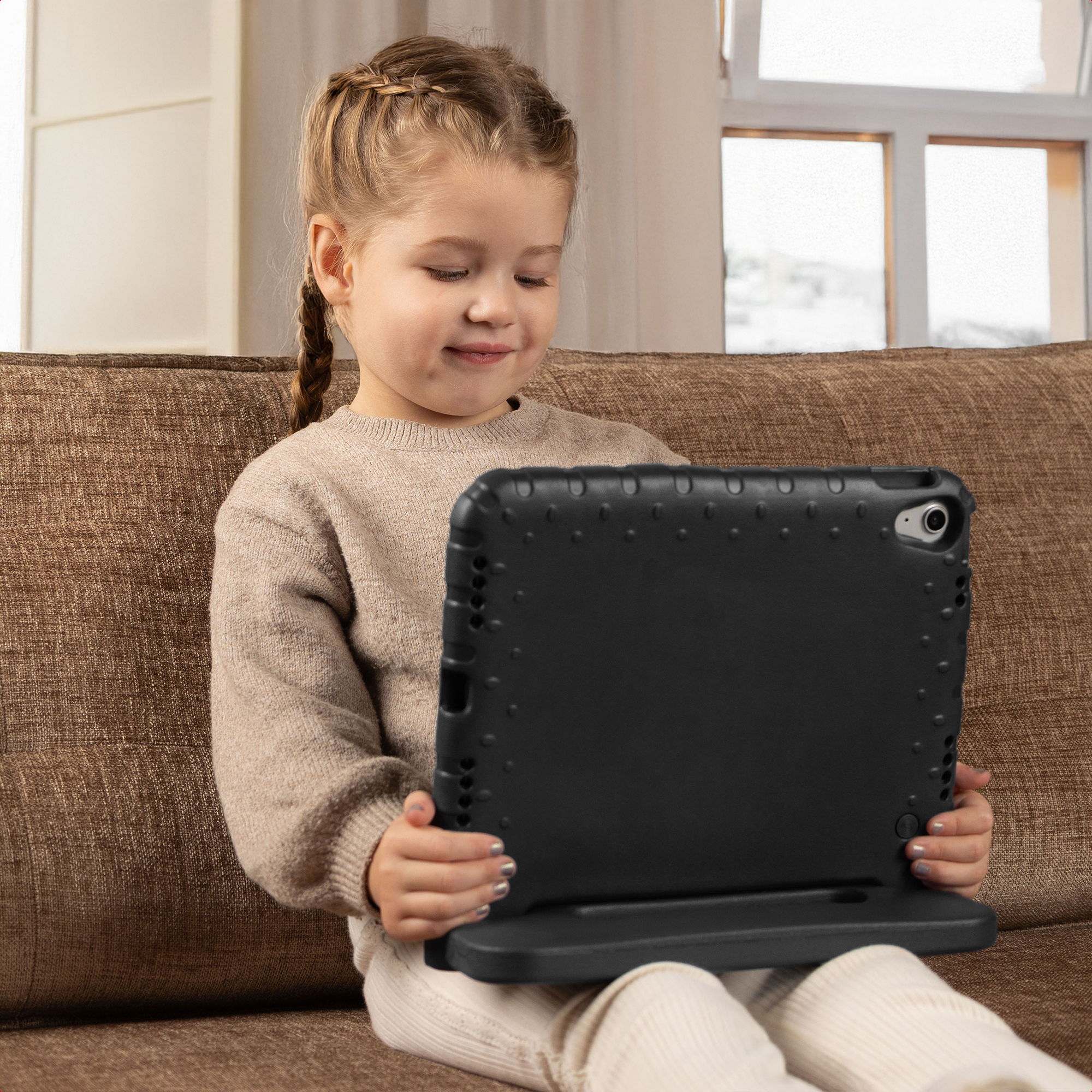 Une jeune fille blonde est assise sur un canapé marron, tenant une tablette dans une coque noire.