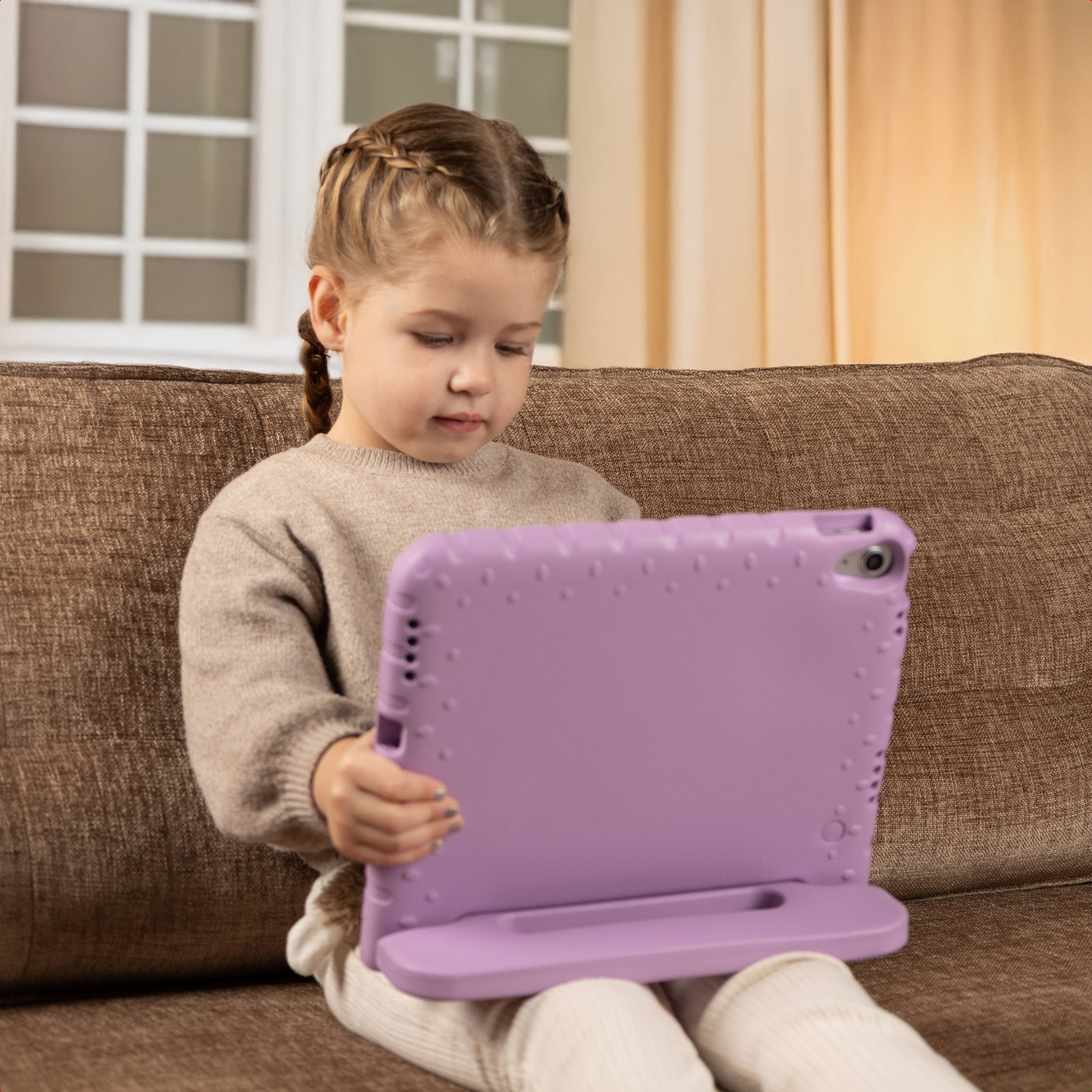 Een jong meisje zit op een bruine bank en houdt een paarse tablet in een standaard vast.