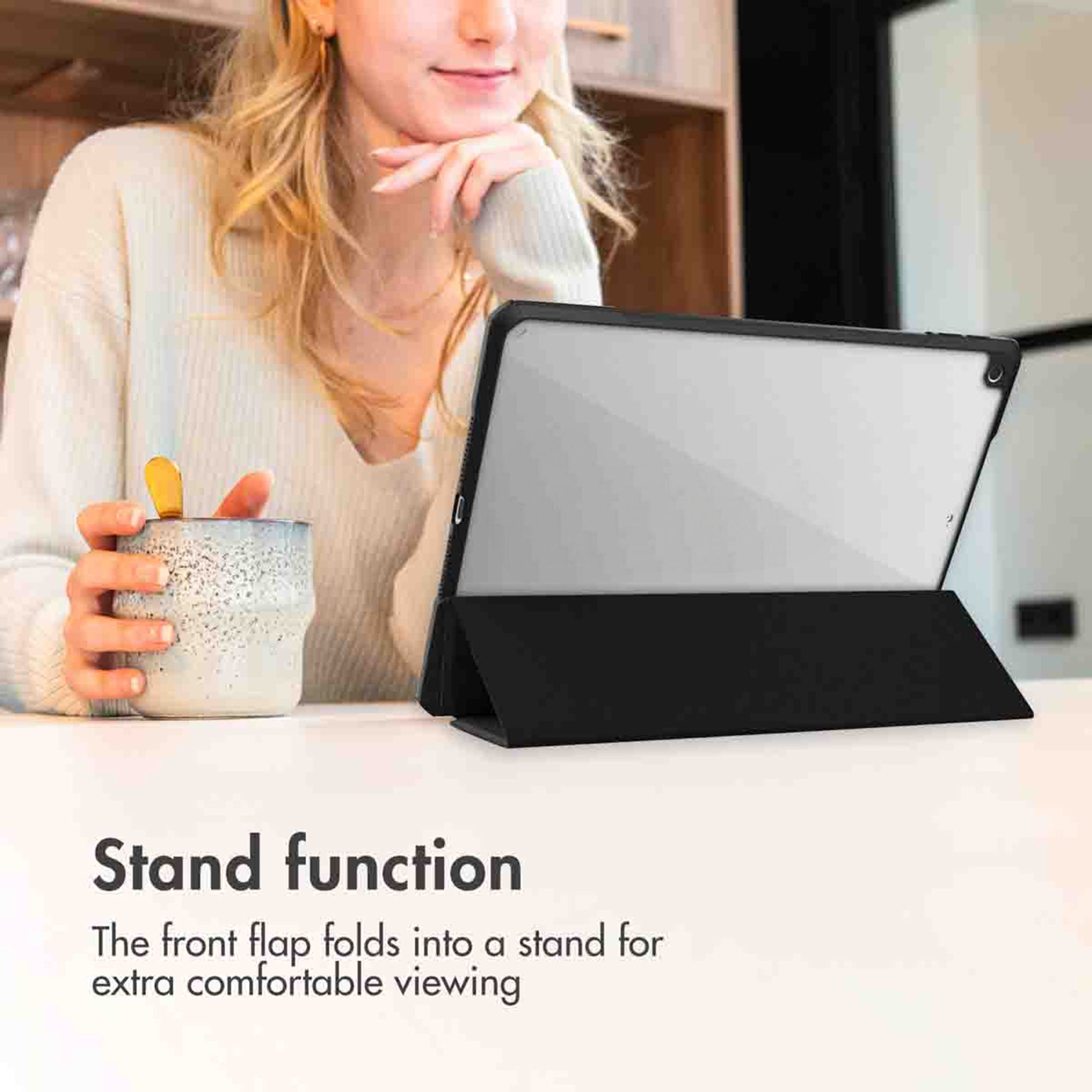 Een blonde vrouw houdt een mok vast naast een tablet in een zwarte standaard op een witte tafel.