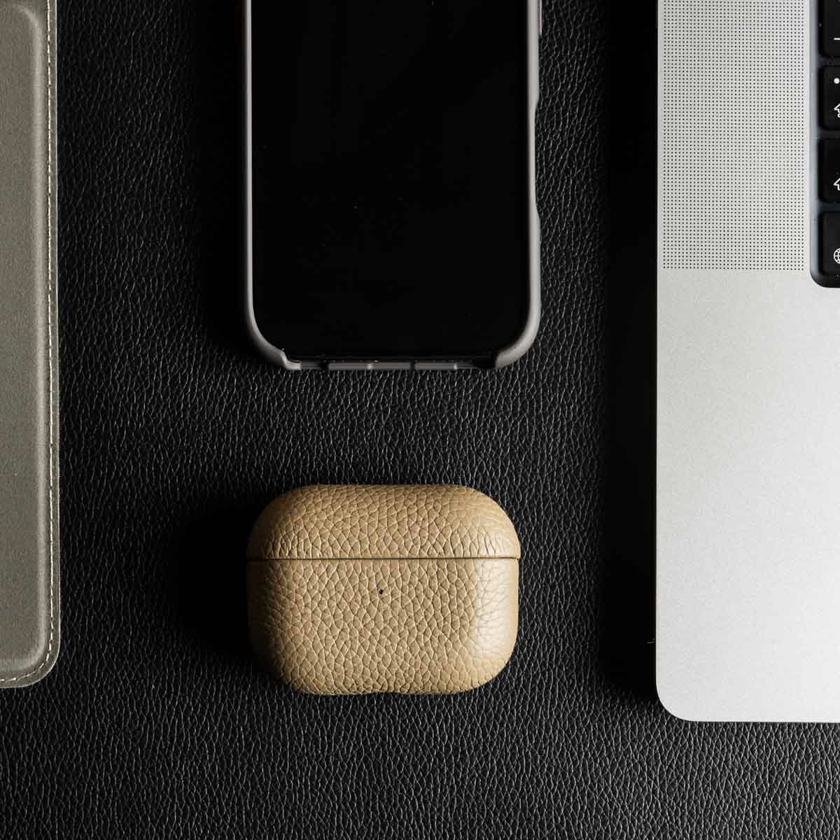 Una custodia in pelle per AirPods si trova su una scrivania in pelle nera, tra uno smartphone e un laptop.