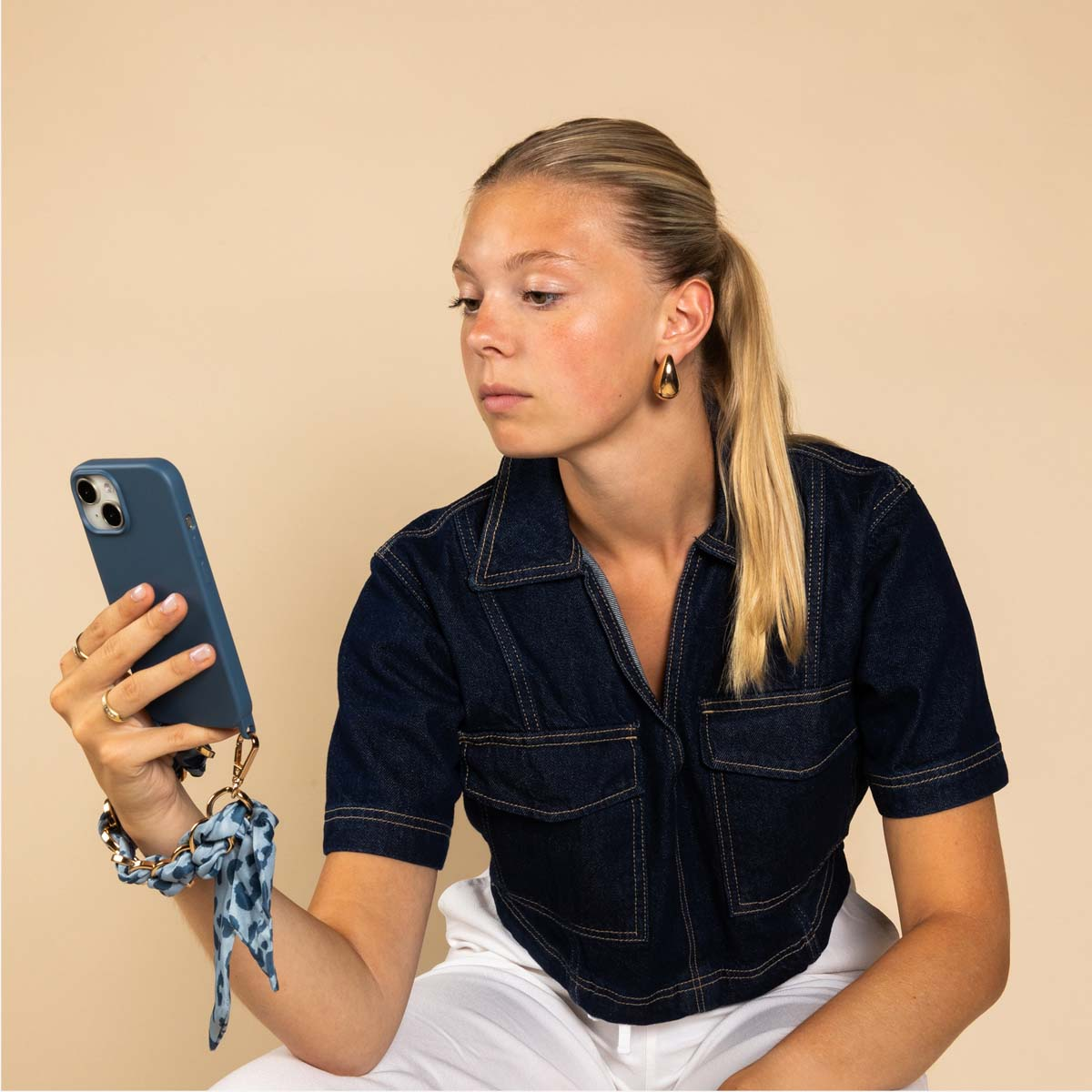 Eine blonde Frau in dunklem Jeanshemd betrachtet ihr blaues Smartphone mit einer gemusterten Handschlaufe.