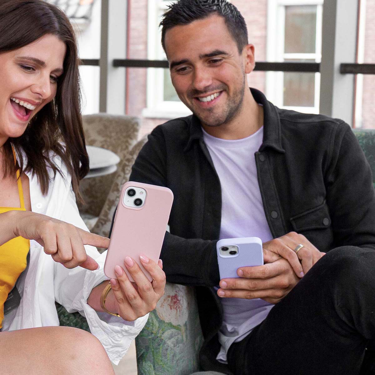 Uśmiechnięta kobieta wskazuje różowy smartfon, a siedzący obok mężczyzna patrzy na swój fioletowy smartfon.