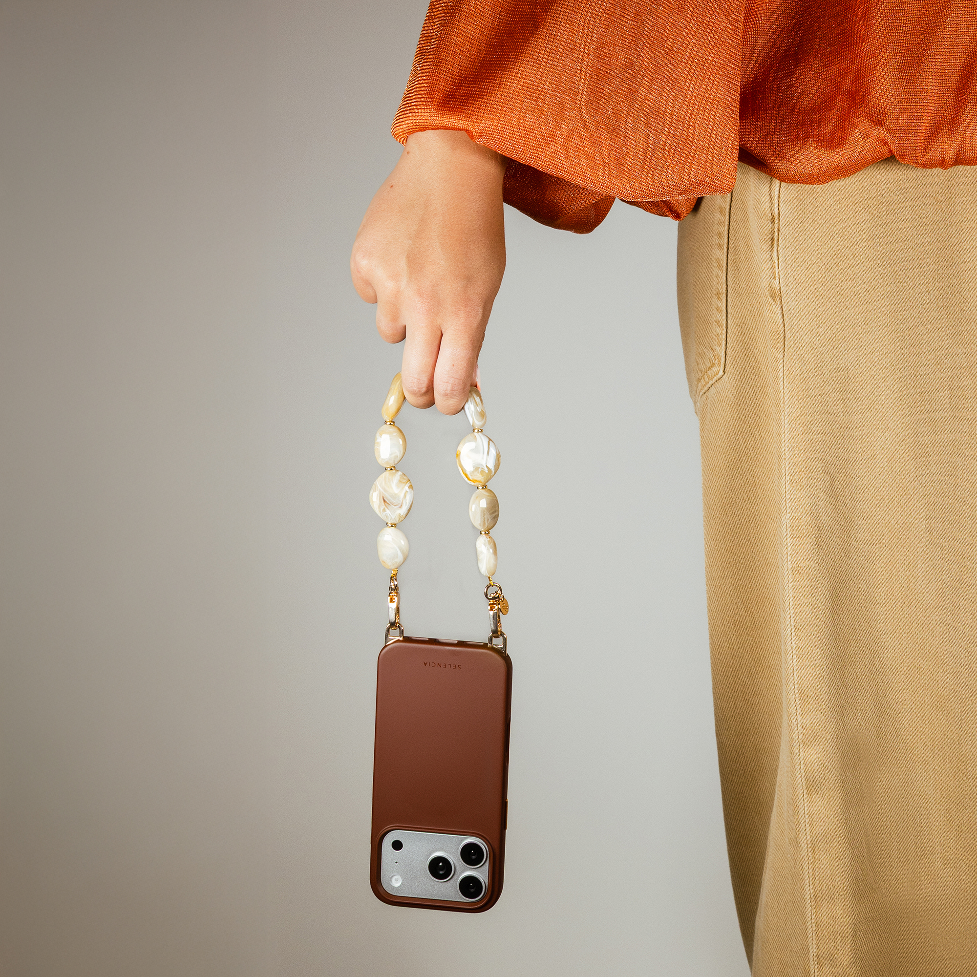 Una persona con top naranja y pantalón beige sostiene una funda de móvil marrón con correa de cuentas.