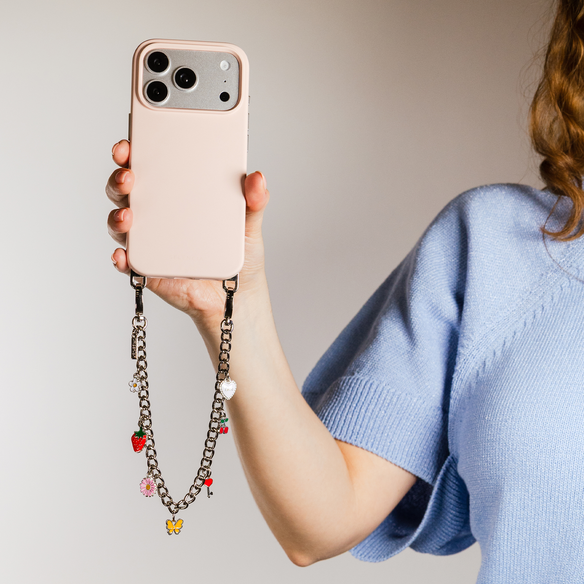 Una persona en jersey azul sostiene un móvil rosa con cadena decorativa.