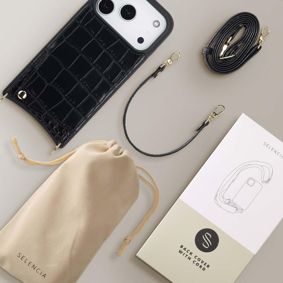 Una funda de móvil negra con patrón de cocodrilo, correas, una bolsa beis y caja sobre superficie neutra.