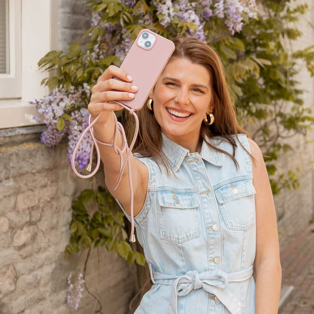 Una mujer sonriente con vestido vaquero se hace un selfi al aire libre con un móvil rosa frente a arbustos en flor.