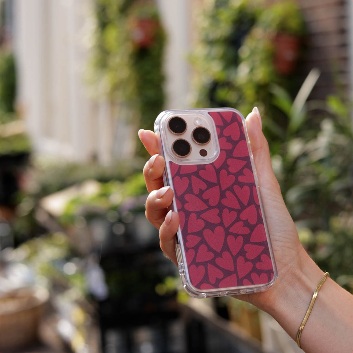 Mano sosteniendo un teléfono con funda de corazones rojos ante un jardín borroso.