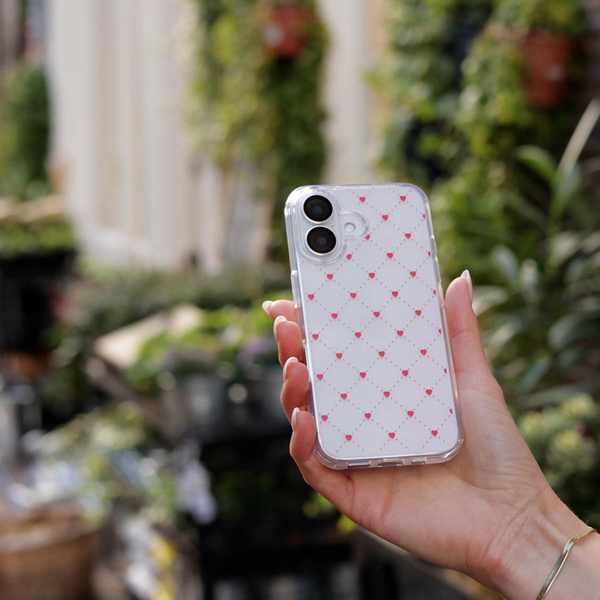 Une main tenant une coque transparente avec motif losange rose et petits cœurs, devant un jardin flou.