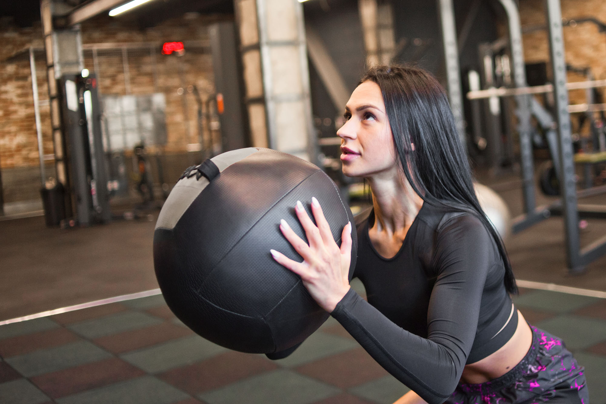 Brunetka w czarnym stroju sportowym trzyma dużą czarną piłkę lekarską na siłowni.