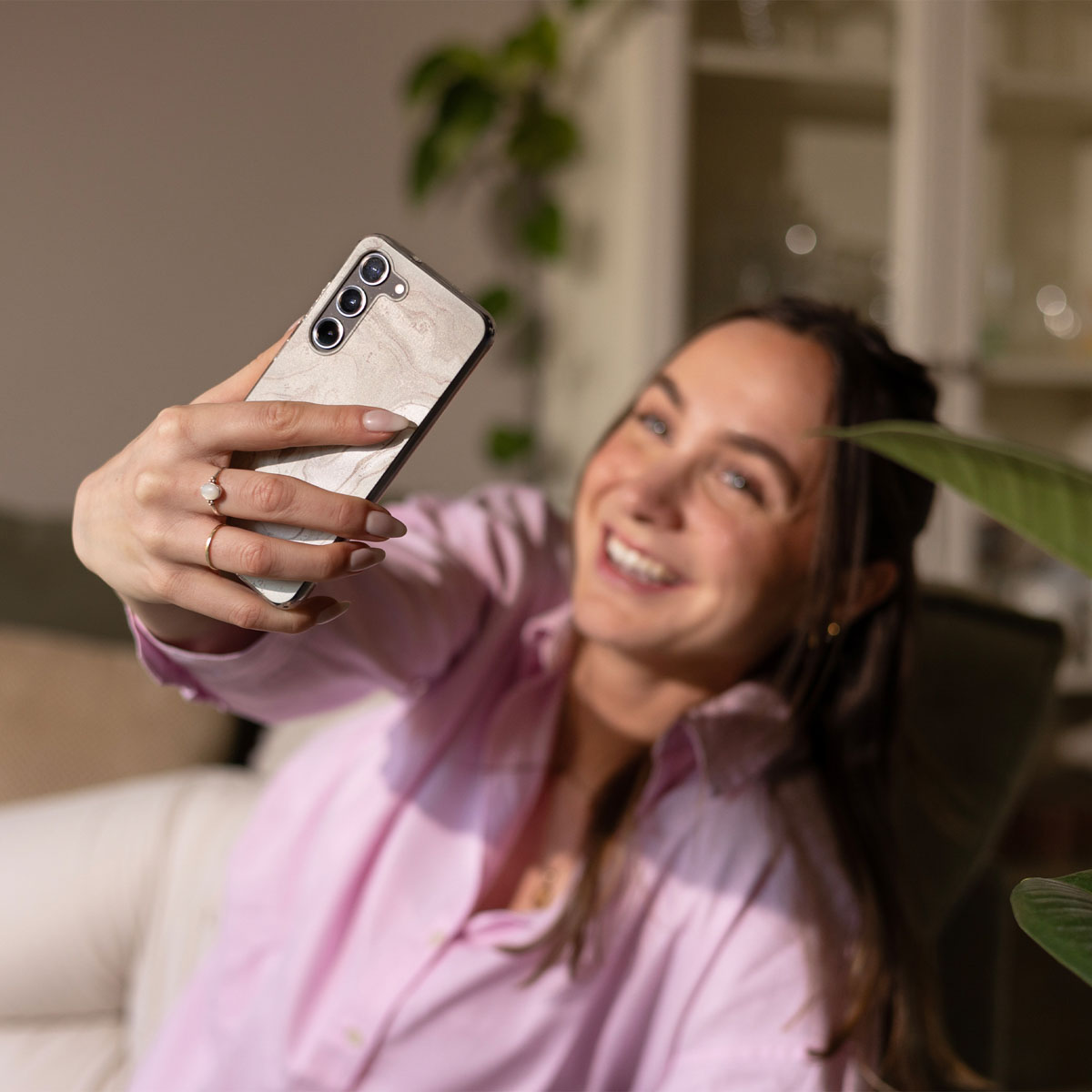 Una mujer sonriente con camisa rosa se hace un selfi con una funda de móvil de mármol en una habitación luminosa.