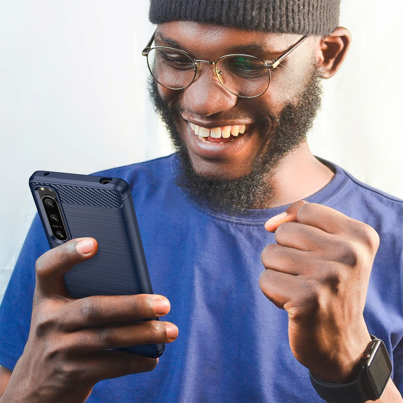 Un uomo barbuto felice con occhiali e maglietta blu tiene un telefono con custodia blu, stringendo il pugno.