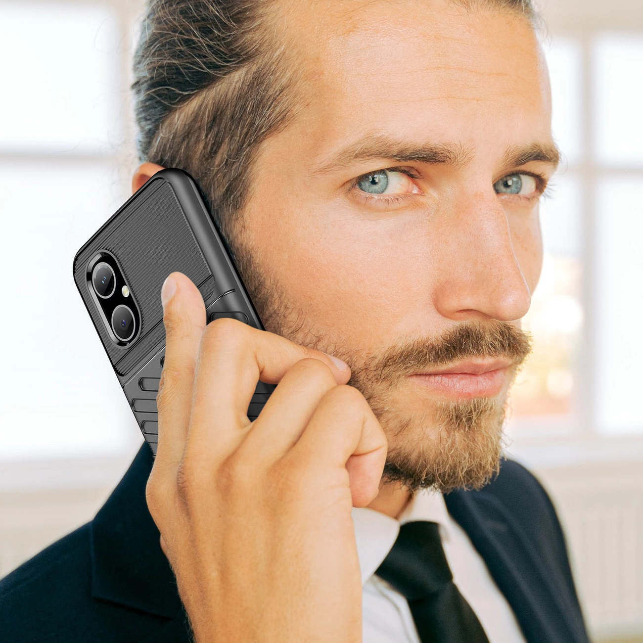 Un uomo barbuto in abito parla al telefono con uno smartphone nero.