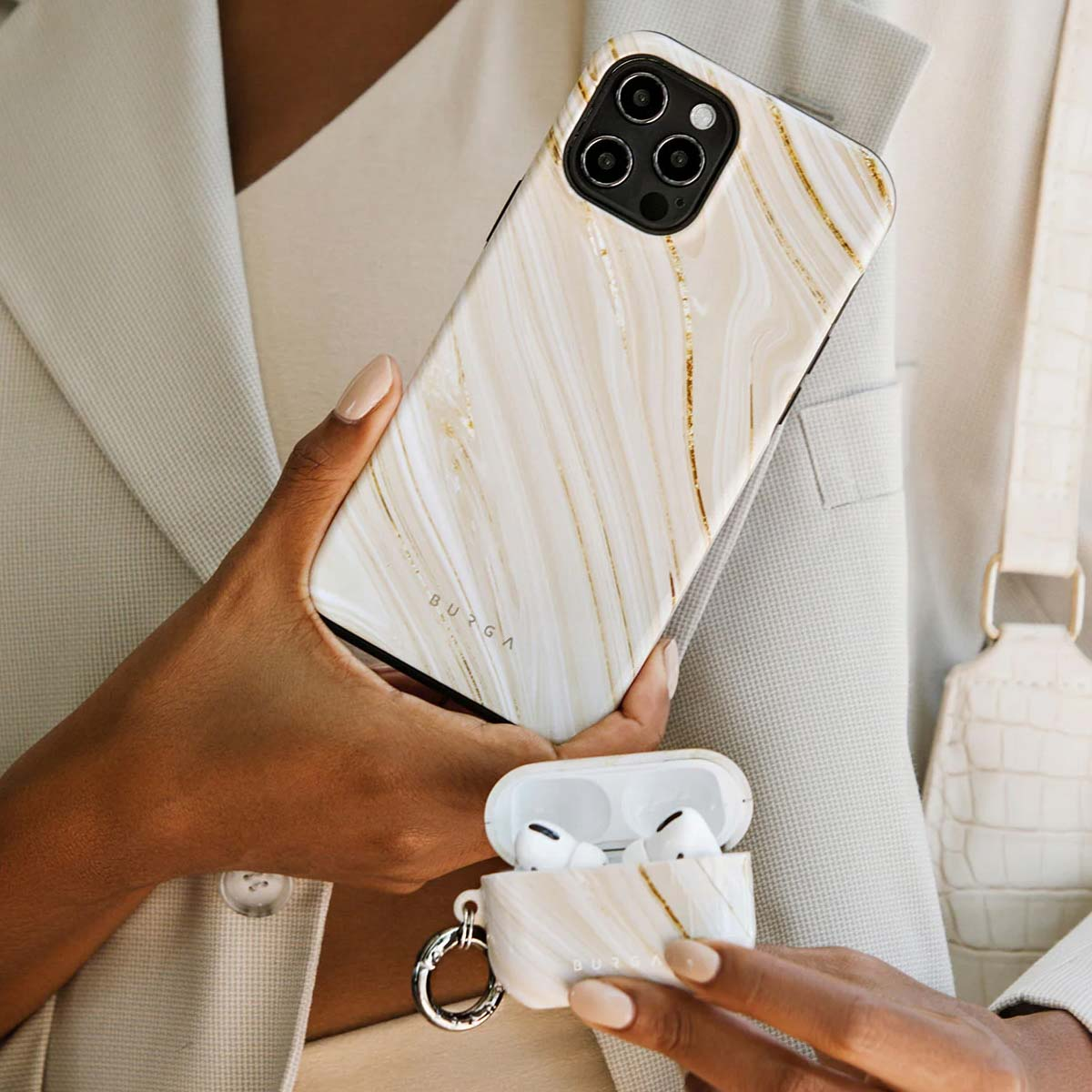 Une personne tient une coque de téléphone et un étui AirPods en marbre blanc et doré, portant un blazer beige.