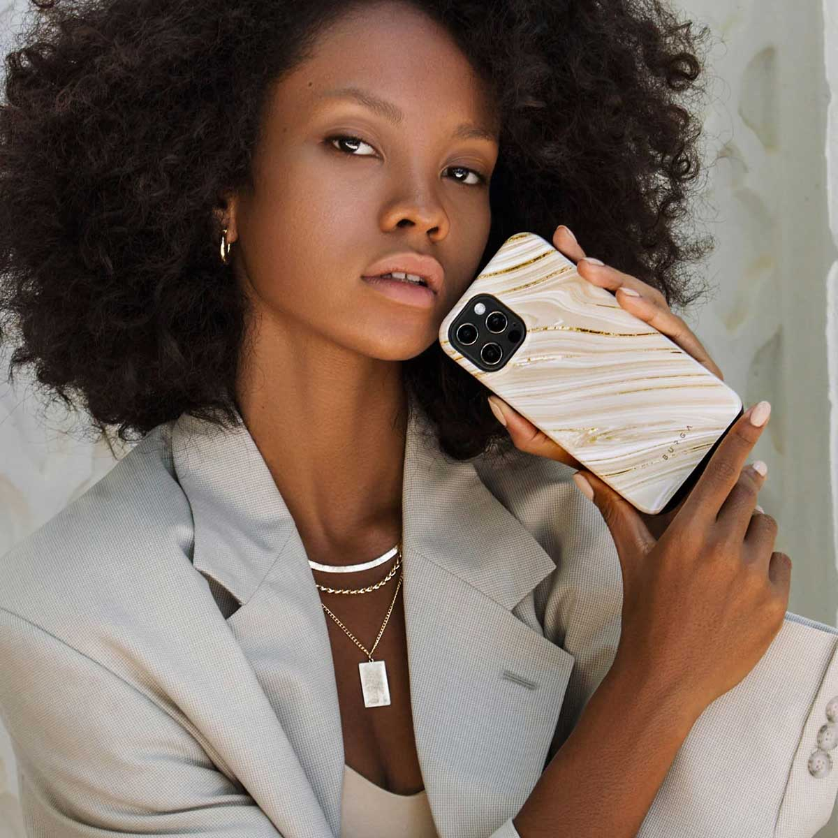 Une femme aux cheveux bouclés foncés et blazer gris tient un smartphone en marbre blanc et doré.