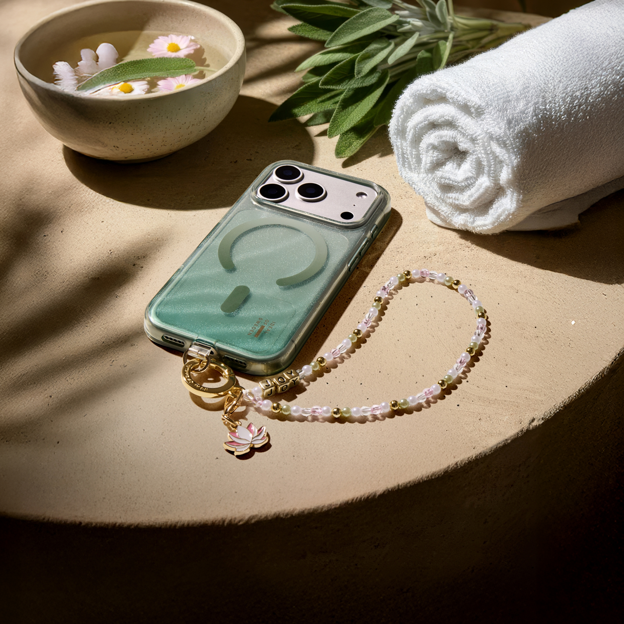 Ein Smartphone mit Perlenkette und Anhänger, eine Wasserschale, grüne Blätter und ein weißes Handtuch auf einer runden Fläche.